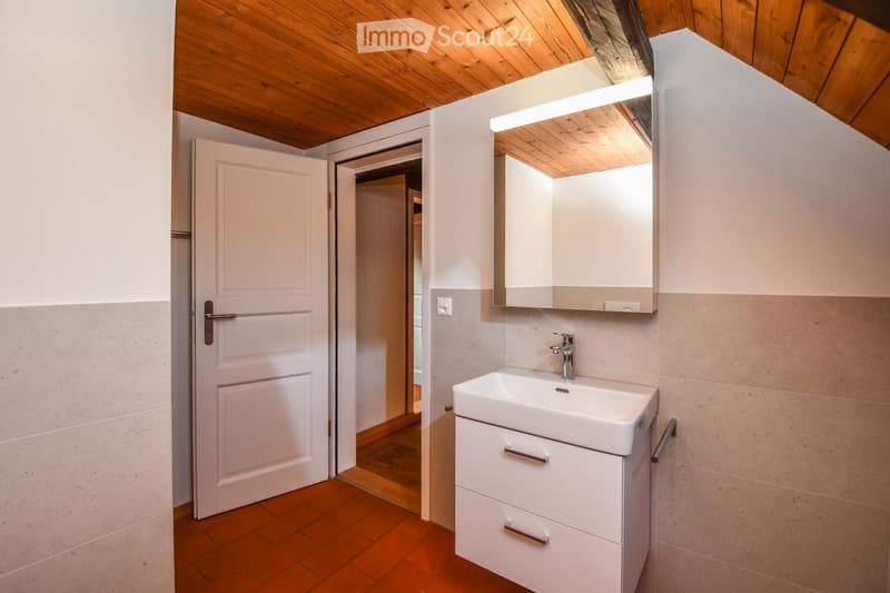 Modern bathroom with a white sink, mirrored cabinet, and wooden ceiling beams. The room features a tiled floor and a door leading to another space.