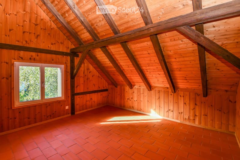 Empty attic space with wooden walls, exposed wooden beams, and a single window letting in natural light. The floor is covered with red brick tiles.