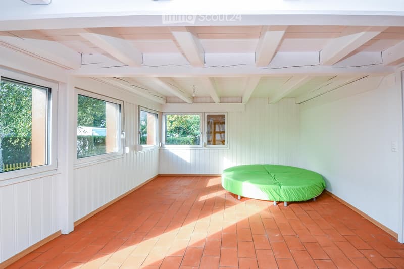 Bright, open room with white wooden paneling, large windows, and a green sofa. The floor is tiled in a warm orange color, and the ceiling has exposed wooden beams.