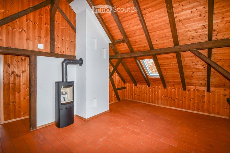 An empty attic space with wooden walls, exposed wooden beams, and a red tiled floor. A modern wood-burning stove is installed against a white wall, with a chimney pipe extending upwards. The room features a skylight window allowing natural light to enter.