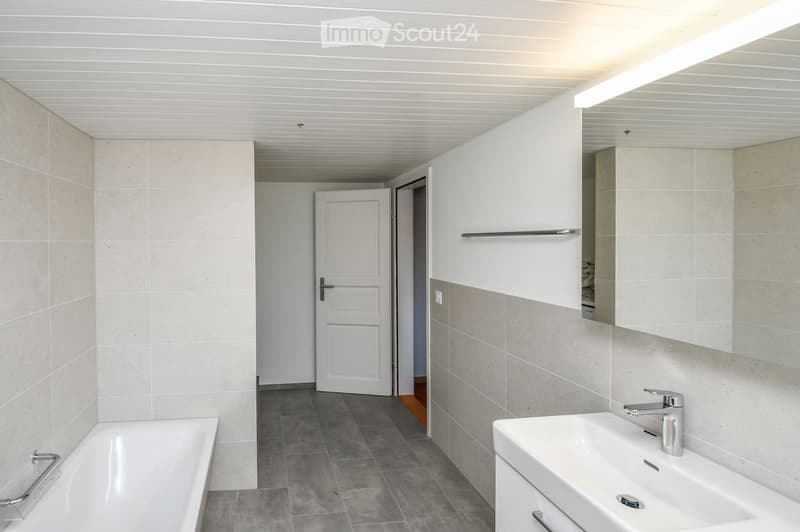 Modern bathroom with a bathtub, sink, mirror, and cabinet. The walls are tiled in a light beige color, and the floor is made of gray tiles. There is a door leading to another room.