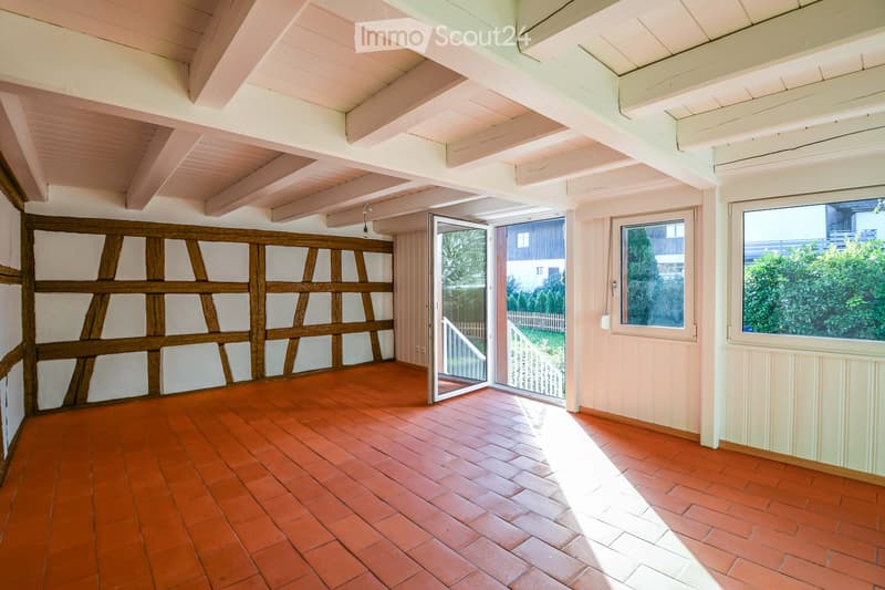 Empty room with exposed wooden beams on the ceiling, red brick floor, large windows, and sliding glass doors leading to an outdoor area.