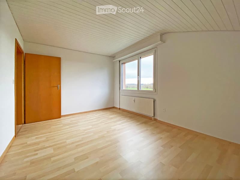 Empty room with light wooden flooring, white walls, a large window with a view, and a radiator below the window. The room has a simple, clean design with no furniture.
