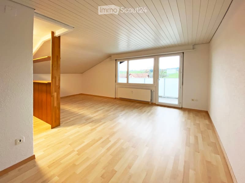 Empty room with wooden flooring, white walls, and a large window with a sliding glass door leading outside. The ceiling has a slanted design typical of an attic space.