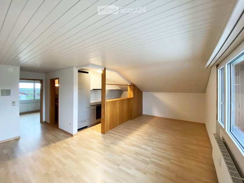 Bright, empty room with wooden flooring and white walls, featuring large windows and a small kitchen area with cabinets and appliances visible in the background.