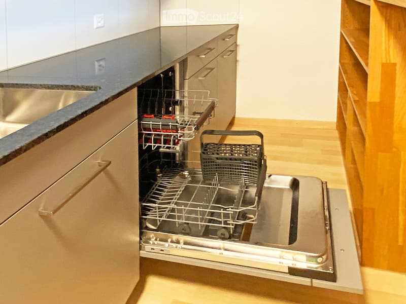 Open stainless steel dishwasher with racks visible, integrated into modern kitchen with wooden flooring and sleek countertops.