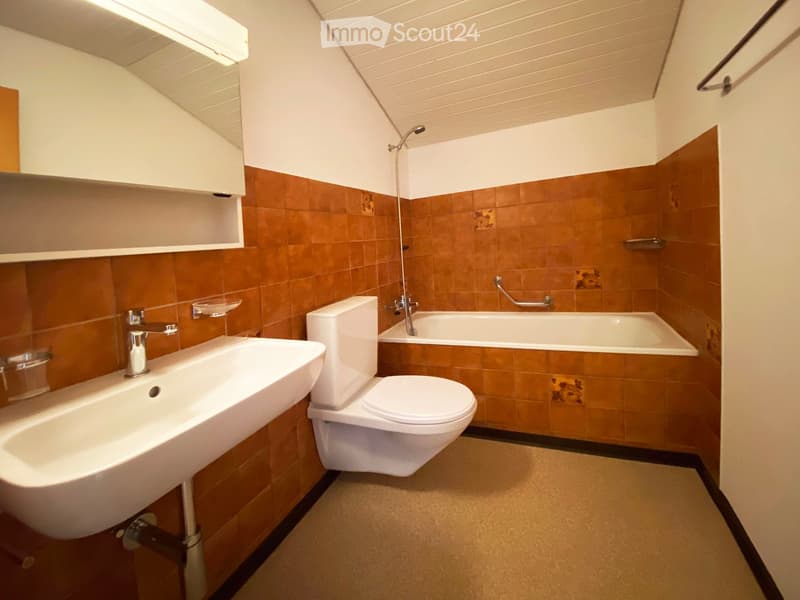 Modern bathroom with a sink, toilet, bathtub, and a mirror above the sink. The walls are tiled in brown and white, and the ceiling has a slanted design.