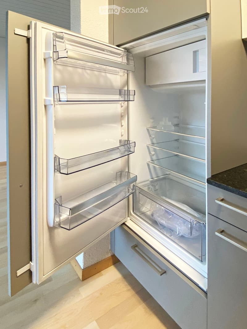 Open refrigerator with multiple shelves and compartments, located in a modern kitchen with light-colored cabinets and a wooden floor.