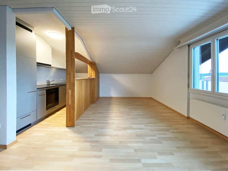 Bright, open room with wooden flooring and white walls, featuring a small kitchen area with cabinets and appliances on the left side and large windows on the right side.