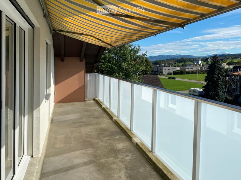 Balcony with a striped awning, modern glass railing, and scenic view of a green landscape and distant mountains.