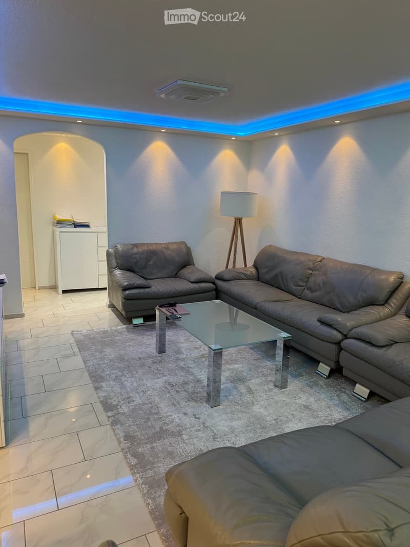 Modern living room with grey leather sofas, a glass coffee table, a floor lamp, and a small white cabinet with books.