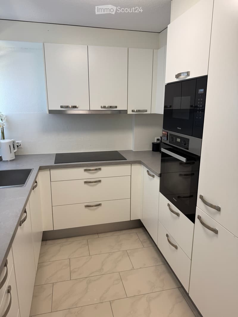 Modern kitchen with white cabinets, stainless steel handles, and grey countertops. Features include a microwave, oven, and electric kettle on the counter.