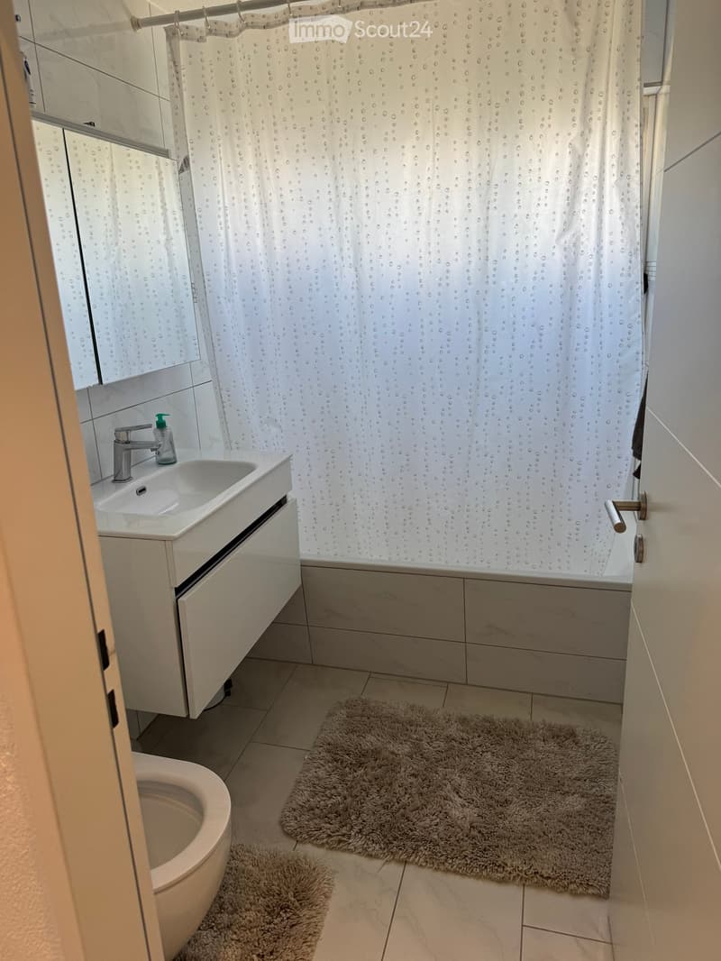 Modern bathroom with a white sink, silver faucet, toilet, bathtub, and a shower curtain with a water droplet pattern. Two plush bath mats are placed on the tiled floor.