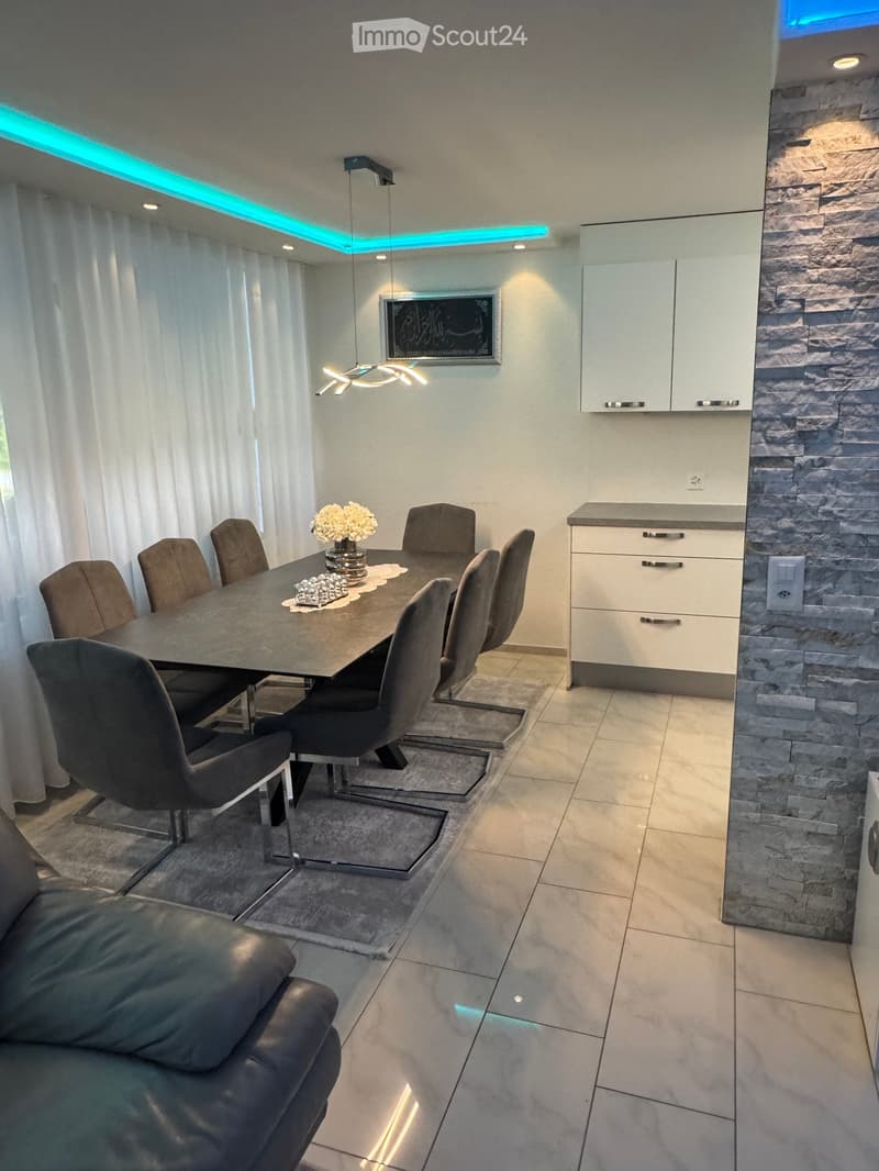 Modern dining and living area with a dark grey dining table, six chairs, a leather sofa, white cabinets, and a decorative flower vase on the table.