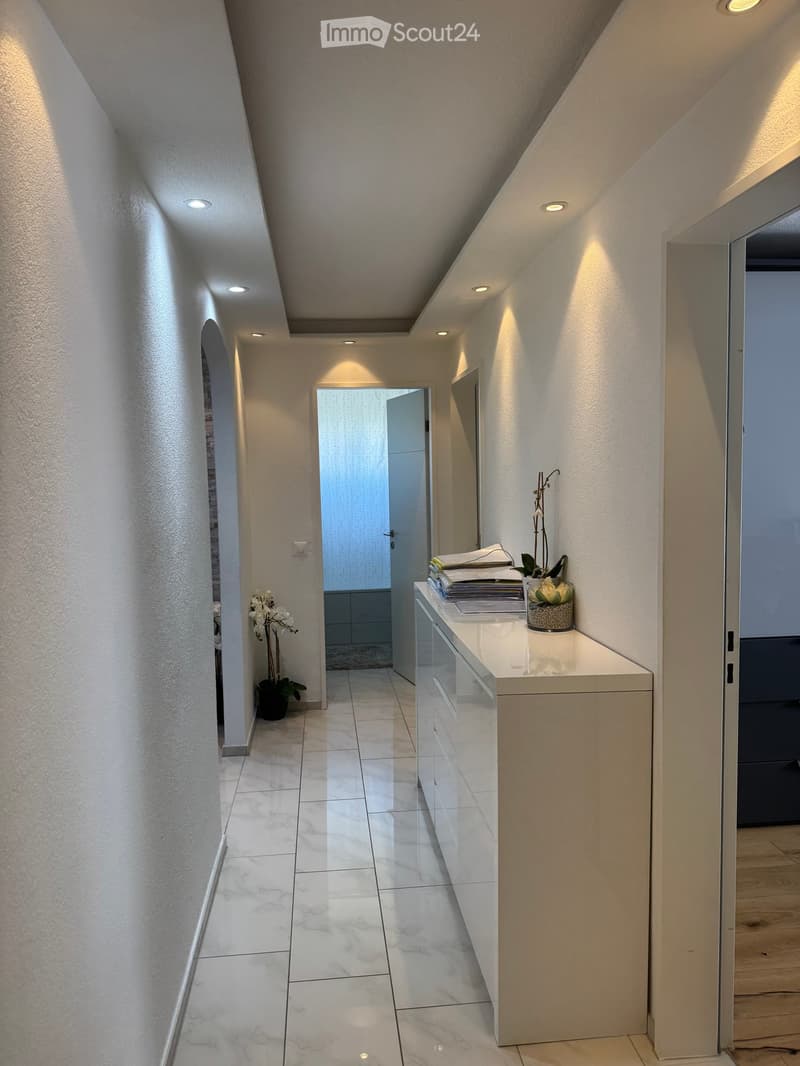 Modern hallway with white marble tiles, recessed lighting, and a small console table with a vase and papers.