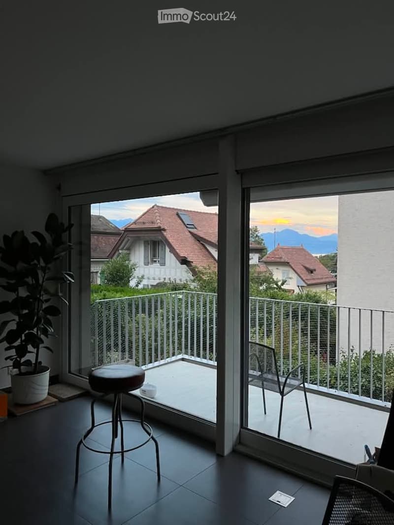 Modern room with large glass doors opening to a balcony, featuring a stool, chair, and potted plant, overlooking a scenic mountain view.