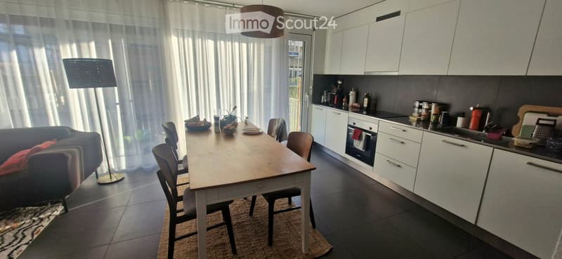 Open-plan living and dining area with a sofa, dining table, chairs, and a modern kitchen featuring white cabinets, an oven, and a sink.