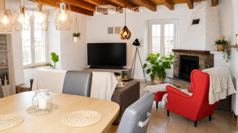 Modern living room with a fireplace, large windows, a sofa, a red armchair, a wooden dining table, and a television. The room features pendant lights and a cozy atmosphere with plants and a candle on the table.