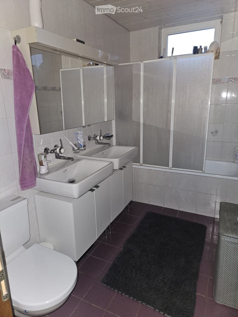 Modern bathroom with two sinks, a toilet, a large mirror, and a shower area with a glass enclosure. The room features a dark-colored rug and a purple towel hanging on the wall.