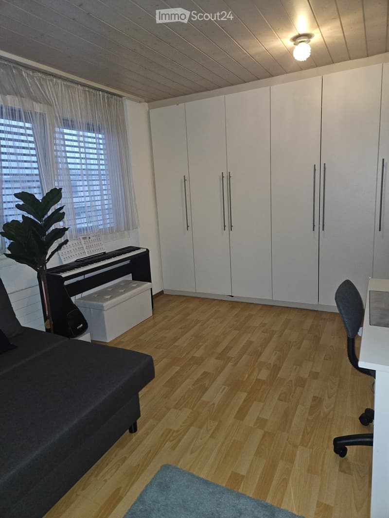 A modern, well-lit room with wooden flooring, white cabinets, a black sofa, a small piano, a blue rug, and a potted plant near the window.