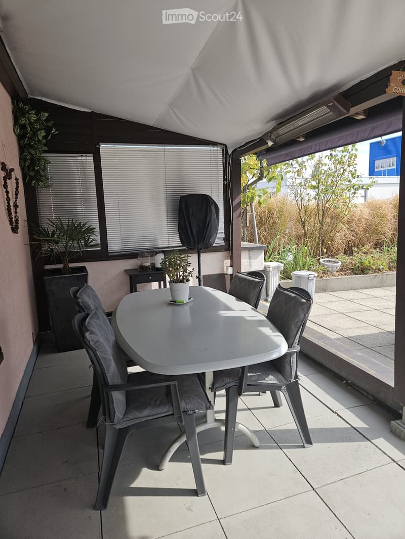 Balcony with a round table, four chairs, and a small potted plant. The area is covered by a retractable awning and has a view of a garden with tall grasses and trees.
