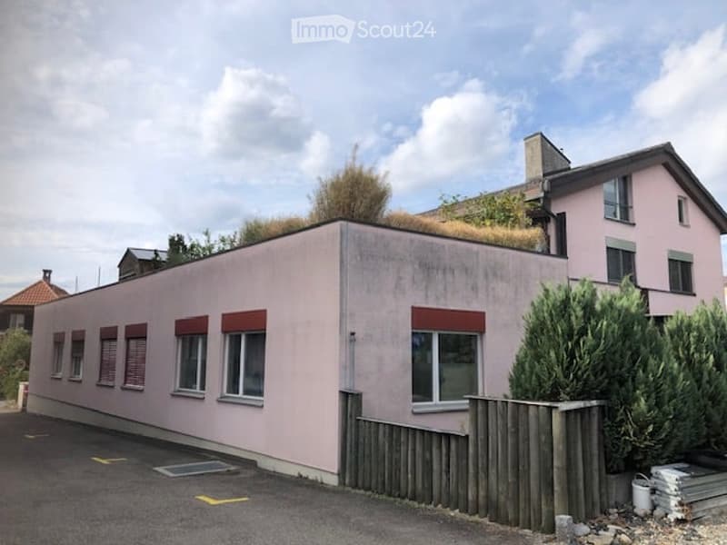 A modern, single-story building with a pink facade, multiple windows with red shutters, and a small green roof. A wooden fence and some greenery are visible in front.
