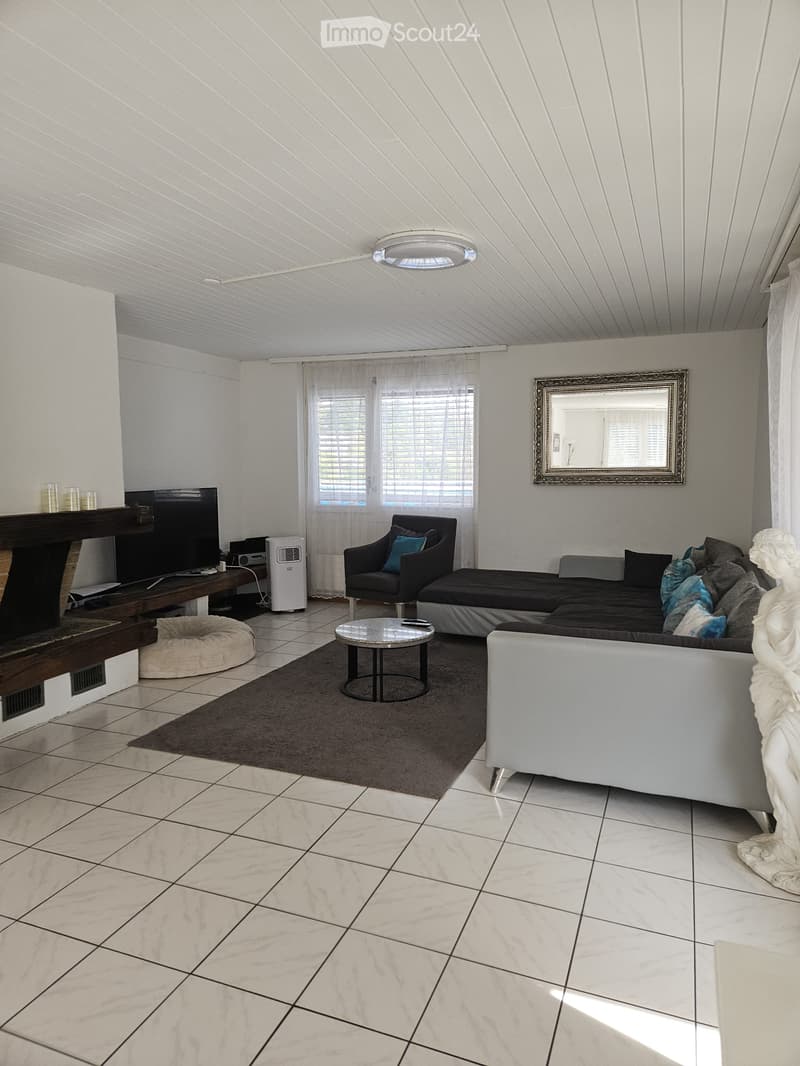 Modern living room with white walls, tiled floor, and large windows. Features a grey sectional sofa, armchair, coffee table, TV, and decorative mirror.