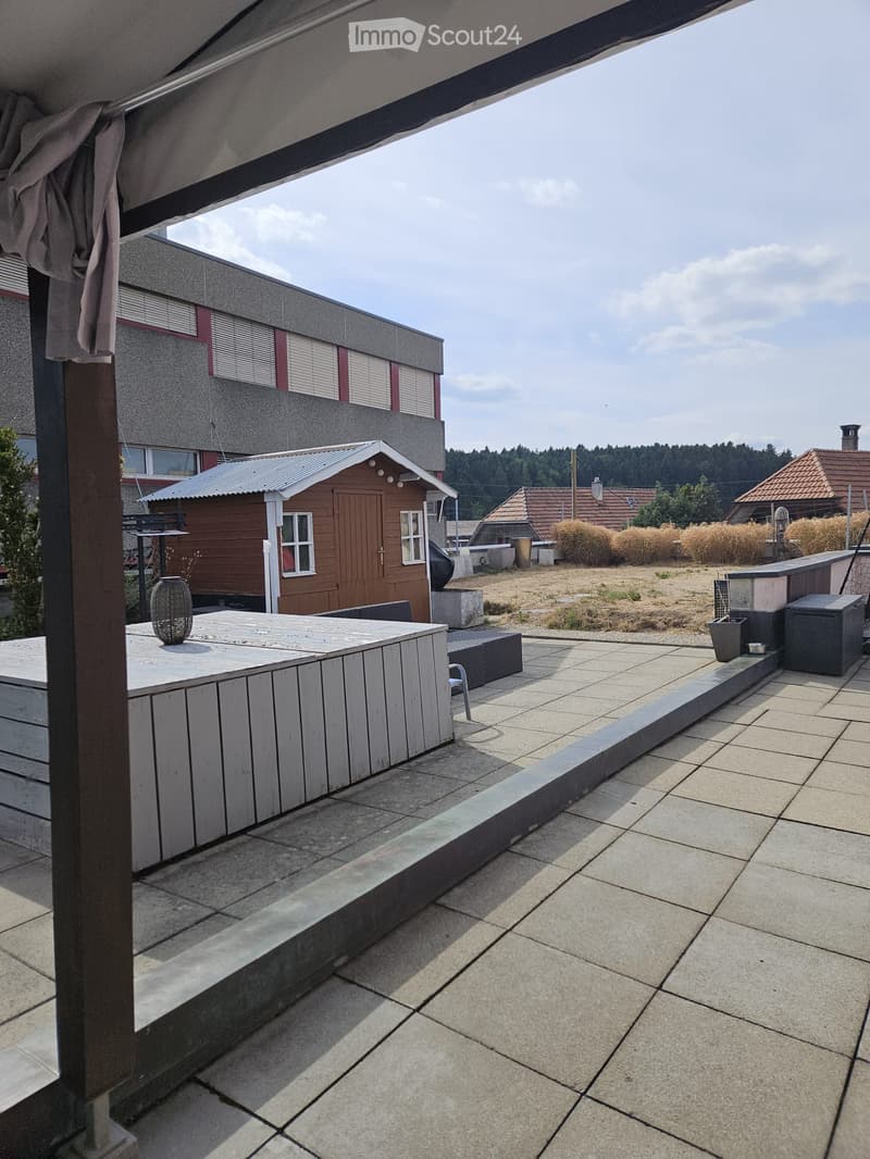 Balcony with tiled floor, wooden table, chair, and decorative vase. Background shows a wooden shed and rural landscape.