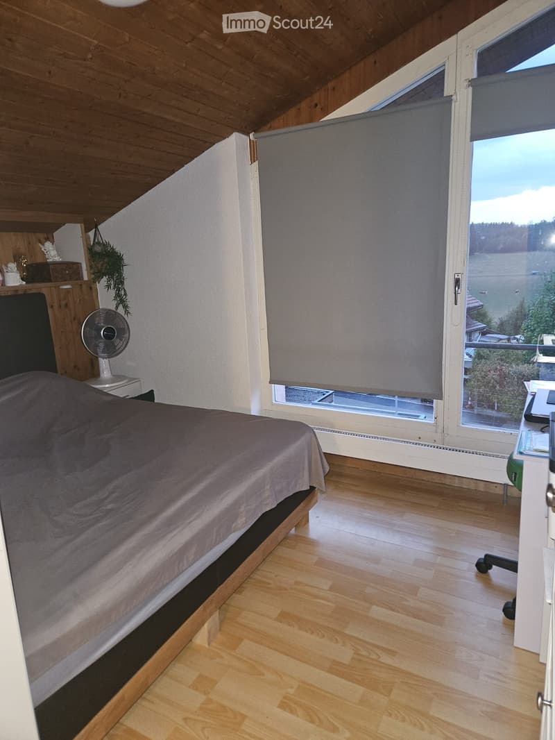 Cozy attic bedroom with wooden slanted ceilings, large windows with blinds, a bed with grey bedding, a small fan, a chair, a table, and a mirror on the wall.