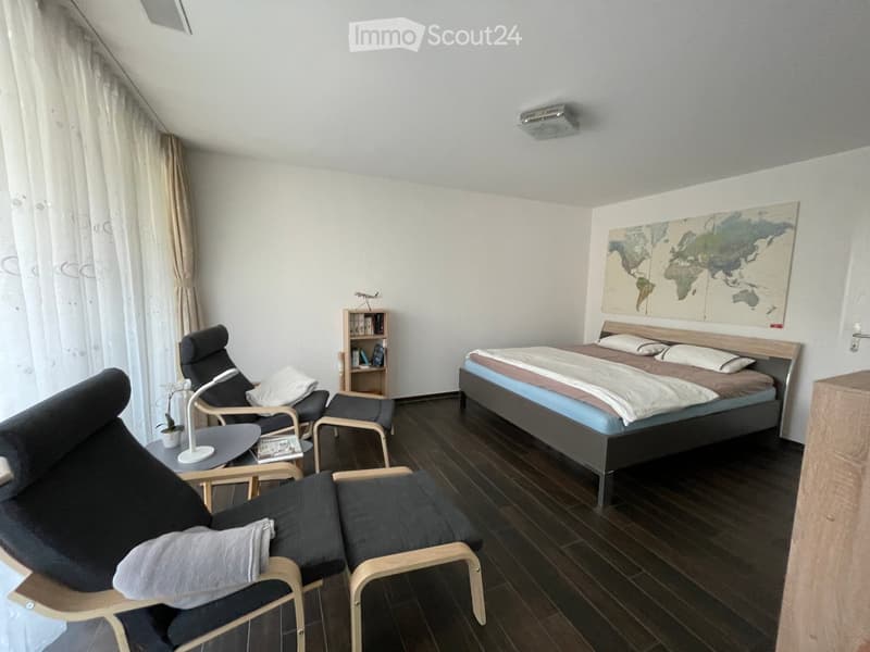 Cozy bedroom with a double bed, two chairs, a small table with a lamp, a bookshelf, and a world map on the wall.