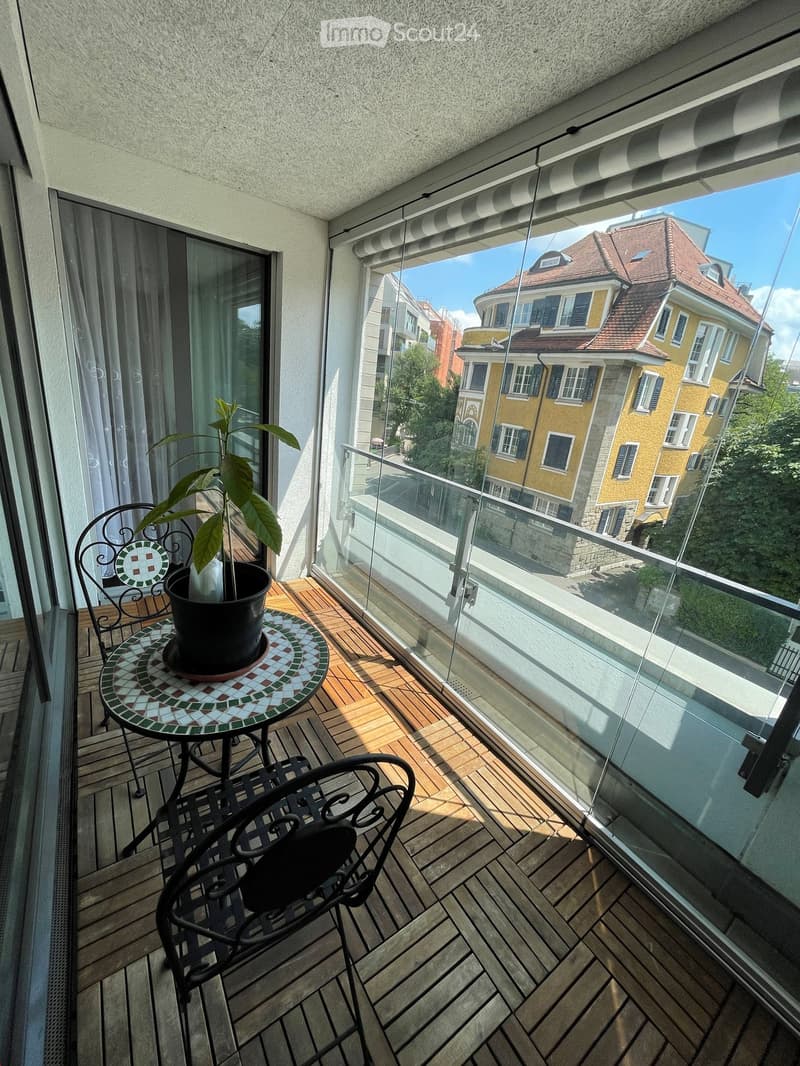 A balcony with wooden floor, glass railing, small round table with a plant, and two chairs
