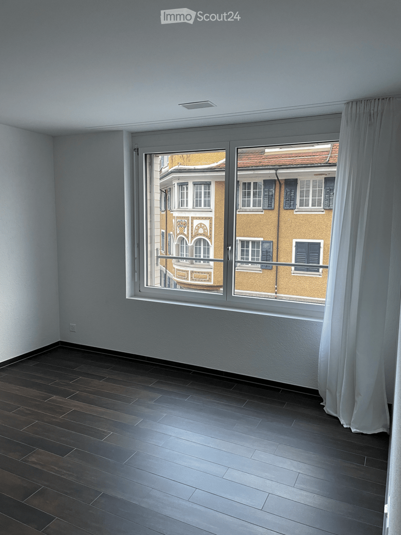Empty room with dark wooden flooring, white walls, and large window with white curtain, offering a view of neighboring buildings.