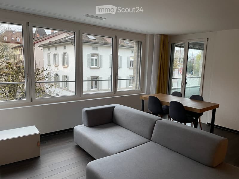 Modern living room with large windows, grey sectional sofa, wooden dining table, and chairs.