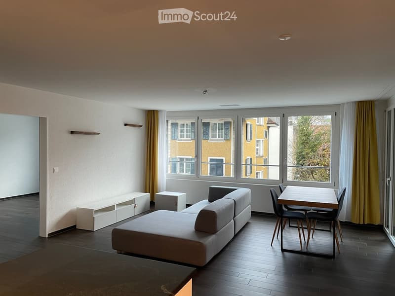Modern living room with dark wooden flooring, large windows, grey sectional sofa, small table with chairs, and white shelving unit.