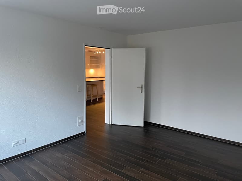 Empty room with dark wooden flooring and white walls, featuring an open doorway leading to a lit kitchen area with visible cabinets and a stool.
