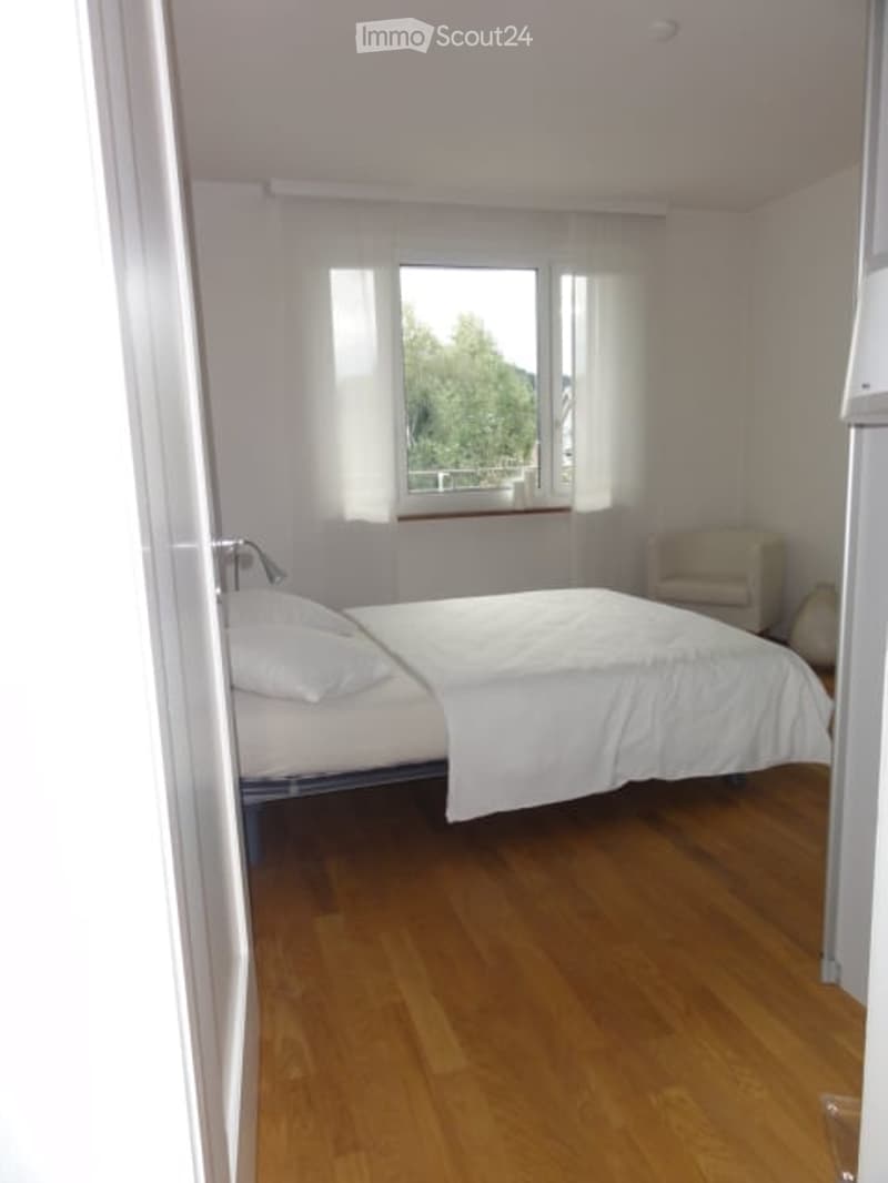 Cozy bedroom with wooden flooring, a neatly made bed with white linens, a small armchair, and large windows with sheer curtains.