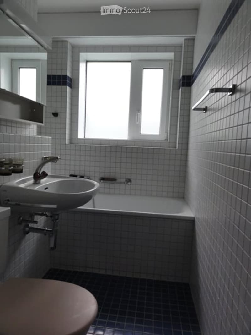 Modern bathroom with white tiled walls and floor, a large window, a sink with a modern faucet, a bathtub, and a toilet. Shelves are mounted on the wall above the sink.