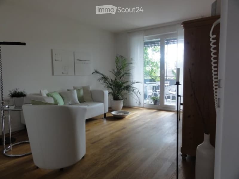 Bright living room with white walls, wooden floor, large windows, and a balcony. Features a white sofa with green and white cushions, a coffee table, an armchair, a floor lamp, a small table with a plant, and a large potted plant near the balcony.
