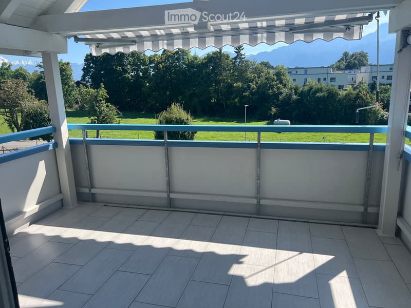 Balcony with tiled floor, white walls, and a striped awning, overlooking a green landscape with trees and mountains.