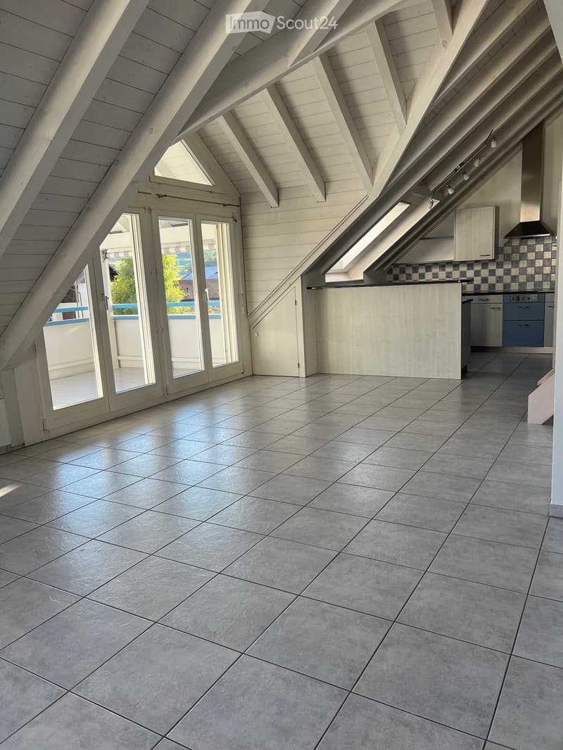 Bright, airy attic space with wooden beams, large skylights, and a modern kitchen area featuring light-colored cabinets and a dishwasher. The floor is tiled, and there is a sliding glass door leading to an outdoor area.