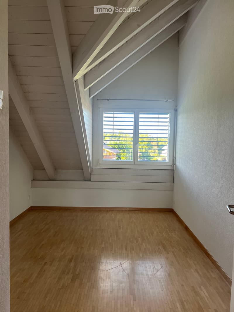 Empty attic room with wooden flooring, white walls, and a skylight window with blinds. The ceiling has exposed wooden beams and a sloped design.
