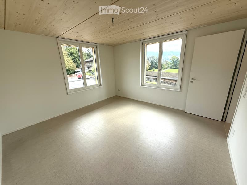 Empty room with light-colored walls, wooden ceiling, and large windows offering a view of the outdoors.