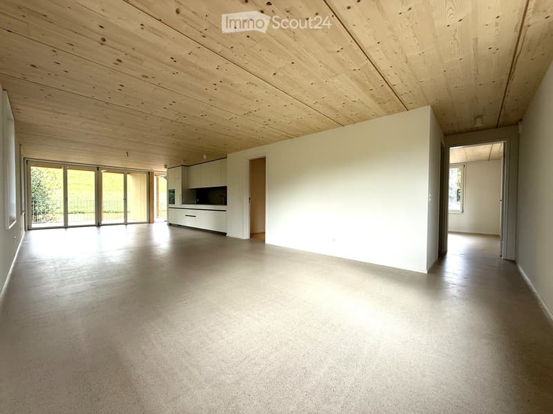 Open-plan unfurnished room with wooden ceiling, concrete floor, white walls, sliding glass doors, and a small kitchen area with cabinets and sink.