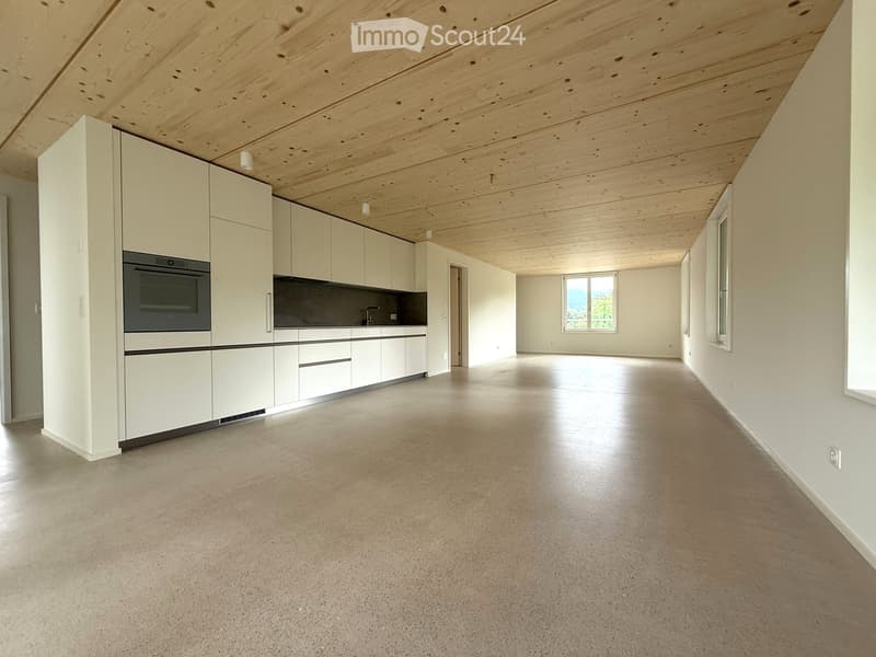 Open-plan space with white cabinetry, built-in oven, and large windows, featuring a smooth concrete floor and exposed wooden ceiling beams.