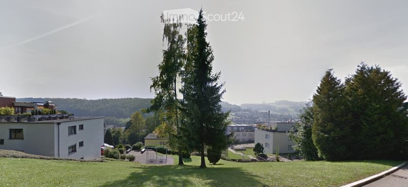 A well-maintained garden area with tall evergreen trees, a grassy lawn, and several modern residential buildings in the background.
