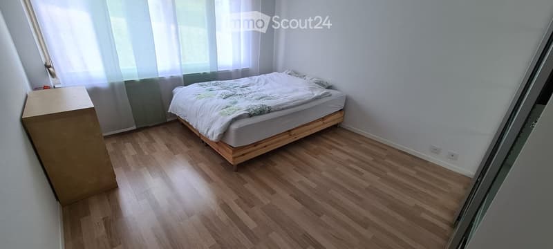 Cozy bedroom with wooden flooring, a single bed with white bedding, and a nightstand. Natural light filters through sheer curtains.