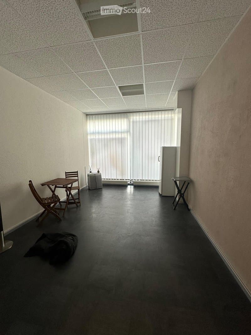 A small, furnished room with a dark floor, a table and chairs, a small refrigerator, and blinds covering a large window.