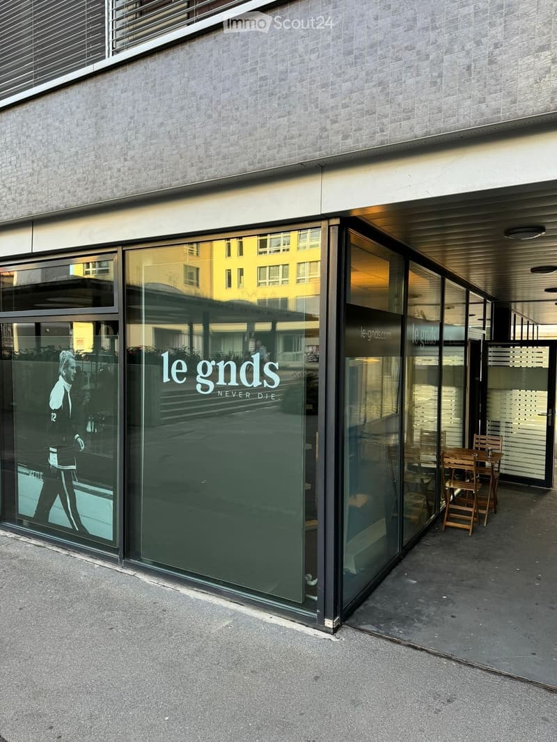 Modern building entrance with large glass windows displaying the text "le gnds NEVER DIE" and a reflection of a person walking. A small outdoor seating area with wooden chairs and a table is visible to the right.