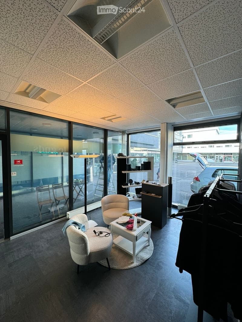 Modern, well-lit room with large windows, featuring two chairs, a small table, a clothing rack, and a shelf with various items.