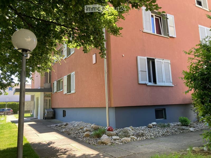 Two-story building with pink facade, white shutters, and a small garden area with rocks and plants.